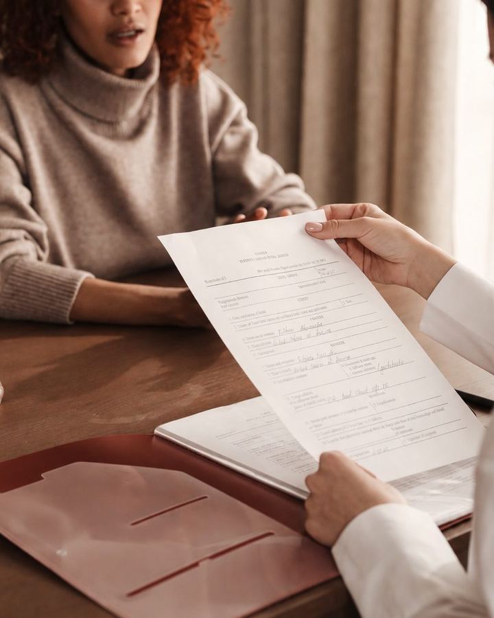 A professional holding a completed form across a table from a client, a folder of papers between them